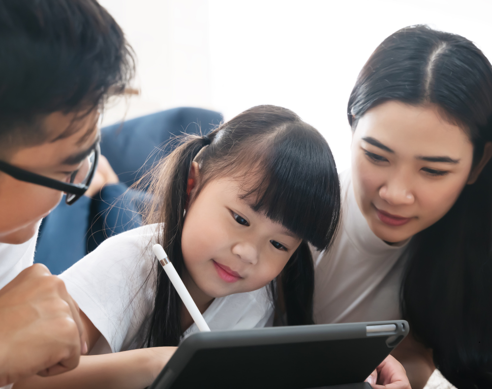 an Asian child drawing on an ipad with her parents next to her watching