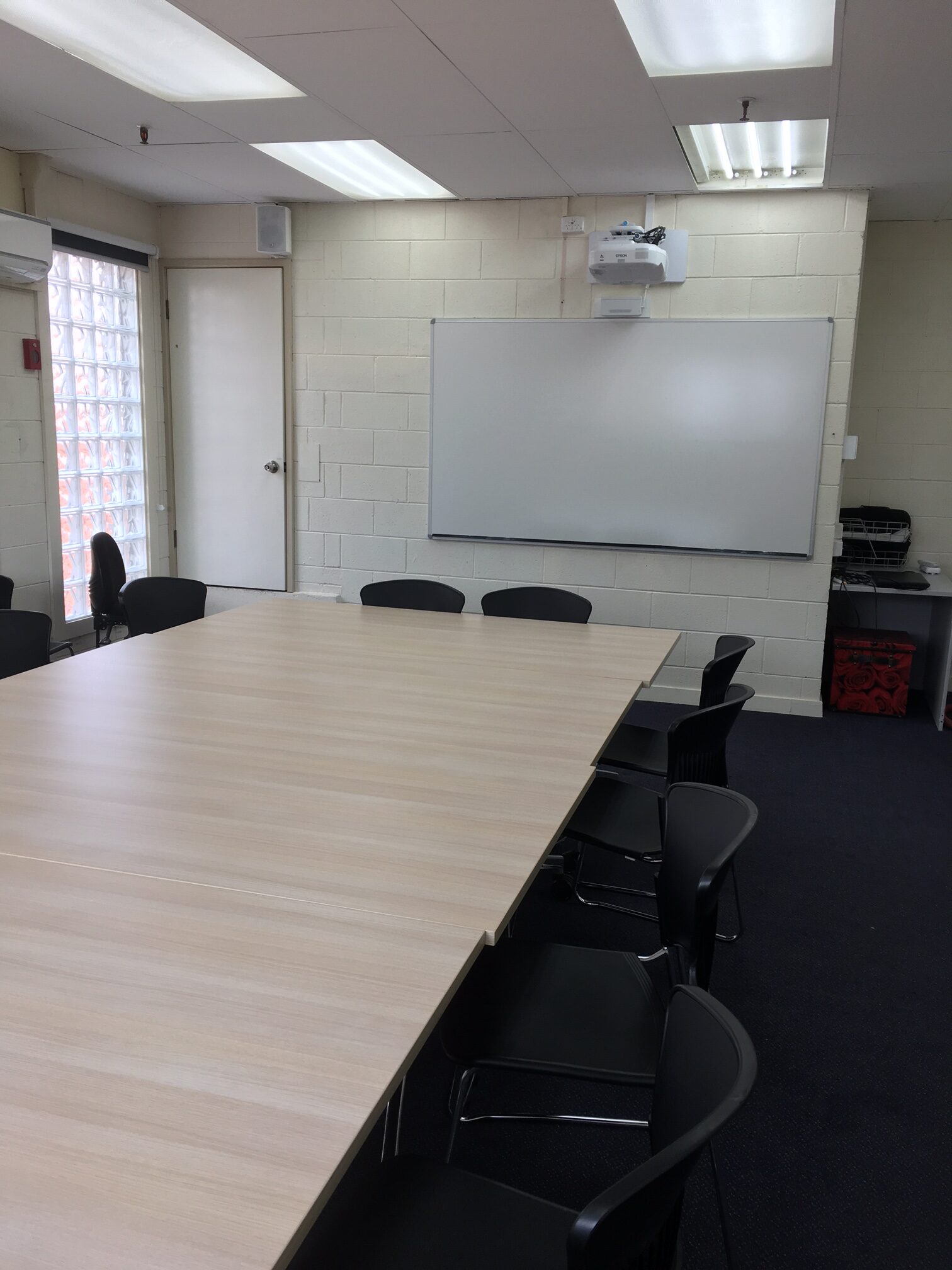 image of a training room with white desks and black chairs around it. a white wall with a whiteboard and projector on ceiling